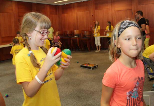 Ein Mädchen mit Zöpfen jongliert mit bunten Bällen in einem Raum voller Kinder.