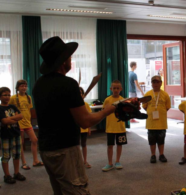 Ein Mann mit Hut erklärt einer Gruppe von Kindern den Umgang mit einer Kettensäge.