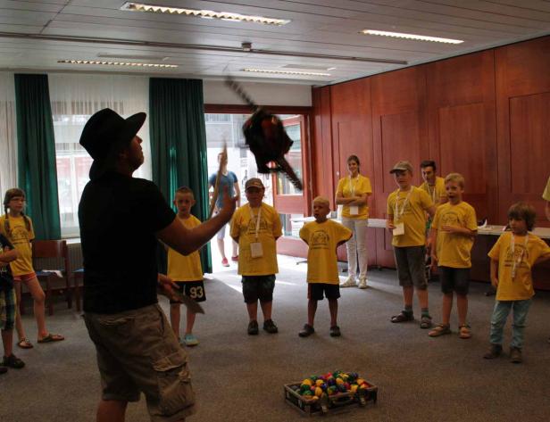 Ein Mann mit Hut jongliert mit einer Kettensäge vor einer Gruppe von Kindern.