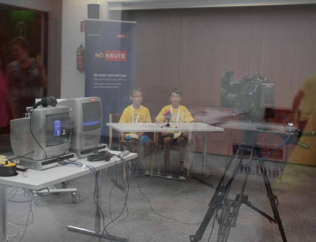 Zwei Jungen sitzen an einem Tisch vor einer Fernsehkamera und Mikrofonen für eine „NÖ Heute“-Sendung.