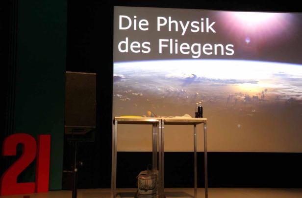 Eine Bühne mit der Aufschrift „Die Physik des Fliegens“ und einem Erdbild im Hintergrund.