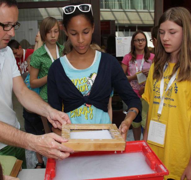 Eine Gruppe von Kindern beobachtet, wie ein Mann einen Papierrahmen in eine Wanne mit Papierbrei taucht.