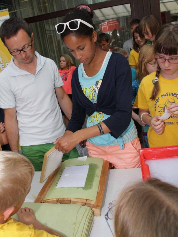 Eine junge Frau schöpft Papier unter Aufsicht eines Mannes, umgeben von Kindern.