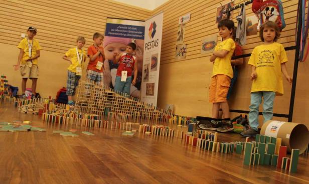 Mehrere Kinder stehen neben einer langen Domino-Kette auf einem Holzboden.