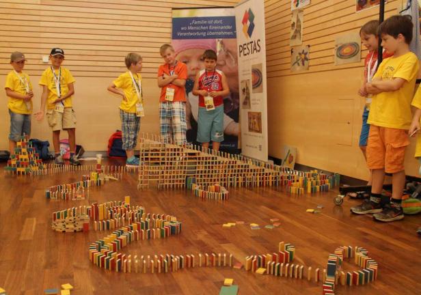 Kinder stehen um eine lange Domino-Kettenreaktion aus Holzbausteinen herum.