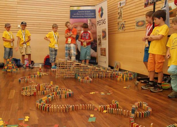 Eine Gruppe Kinder betrachtet eine Strecke aus Dominosteinen und Holzklötzen auf einem Holzboden.