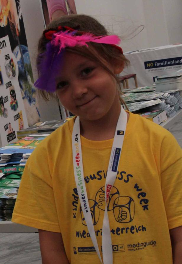 Ein Mädchen mit Federn im Haar trägt ein gelbes T-Shirt mit der Aufschrift „Kinder Business Week“.