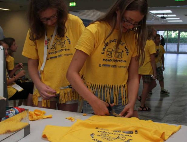 Zwei Mädchen schneiden Fransen in gelbe T-Shirts mit dem Aufdruck „Kinder Business Week Niederösterreich“.