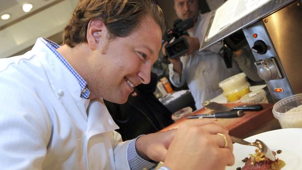 Ein lächelnder Koch garniert ein Gericht mit einem Löffel, während er gefilmt wird.