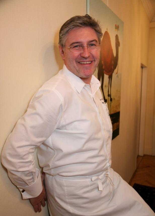 Ein lächelnder Mann in Kochkleidung lehnt an einer Wand mit einem Kamelbild im Hintergrund.