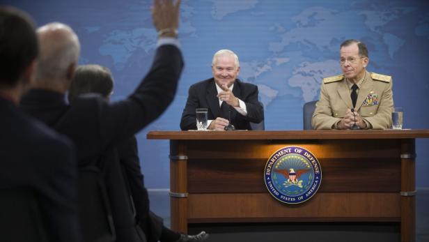 Zwei Männer sitzen an einem Tisch mit dem Siegel des US-Verteidigungsministeriums vor einer Weltkarte.