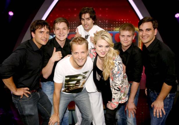 Eine Gruppe von sieben Personen posiert lächelnd vor einem roten Hintergrund.