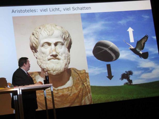 Ein Mann hält eine Präsentation mit einer Abbildung von Aristoteles und einem Stein, der auf einen Baum fällt, während eine Taube aufsteigt.