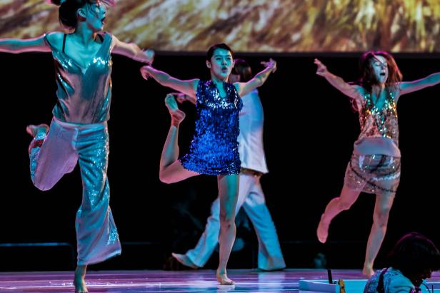 Tänzerinnen in glitzernden Kostümen führen eine Choreografie auf einer Bühne auf.