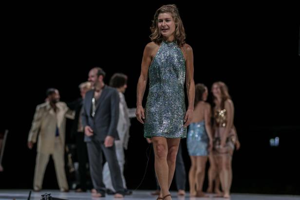 Eine Frau in einem glitzernden Kleid steht auf einer Bühne vor anderen Darstellern.