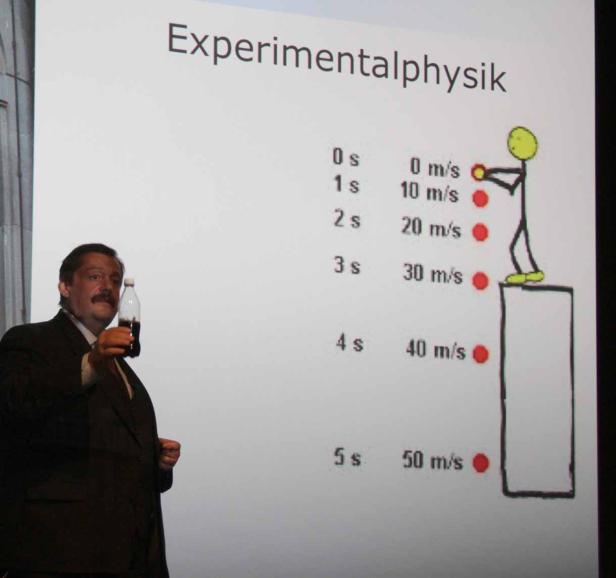 Ein Mann im Anzug präsentiert eine Flasche vor einer Tafel zum Thema Experimentalphysik.