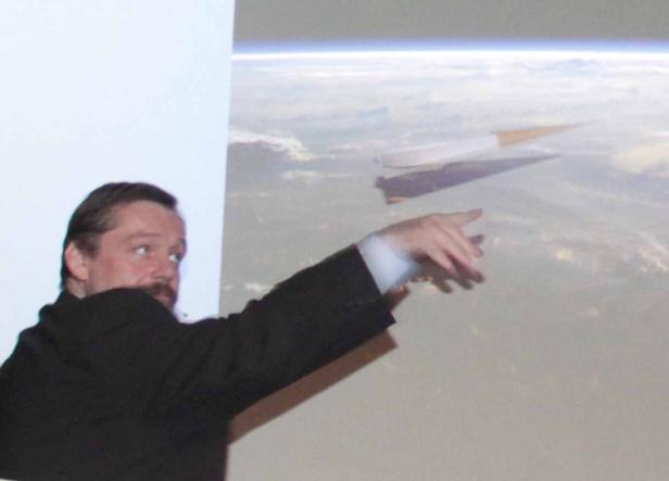 Ein Mann präsentiert ein Raumfahrzeug vor einem Hintergrund mit Wolken und Erdkrümmung.