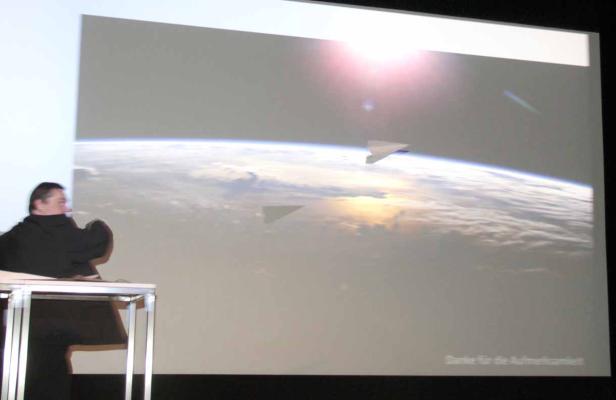 Eine Person hält einen Vortrag mit einer Projektion der Erde und Papierflugzeugen.