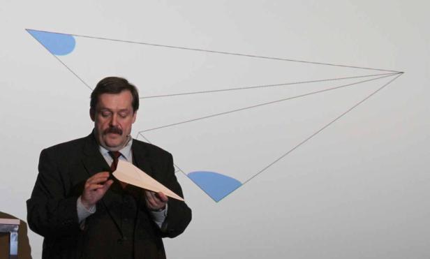 Ein Mann im Anzug hält ein Papierflugzeug vor einer Präsentation mit geometrischen Formen.