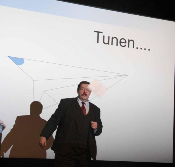 Ein Mann im Anzug präsentiert eine Folie mit der Aufschrift „Tunen…“.