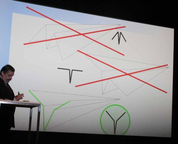 Ein Mann steht vor einer Präsentation mit Papierflieger-Skizzen, die teilweise durchgestrichen sind.