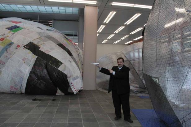 Ein Mann im Anzug steht zwischen zwei großen, kugelförmigen Kunstobjekten und hält ein Papierflugzeug.