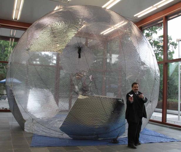 Ein Mann im Anzug steht neben einer großen, silbernen Kugel und hält einen Papierflieger.