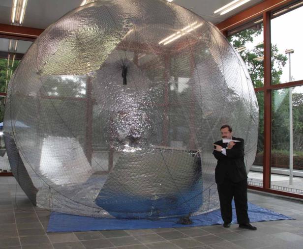 Ein Mann im Anzug steht vor einer großen, netzartigen Kugel in einem Raum.