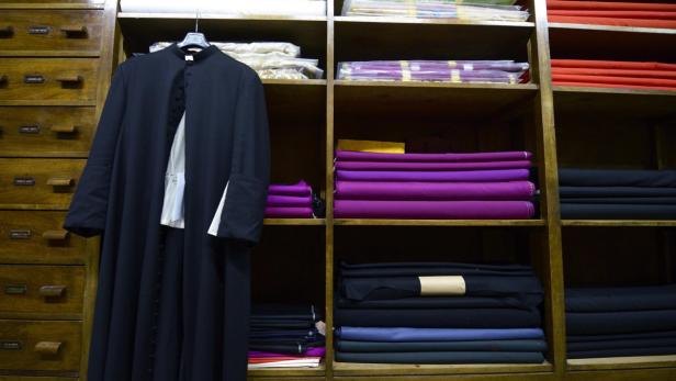 Eine Soutane hängt in einem Schrank neben Stoffballen in verschiedenen Farben.