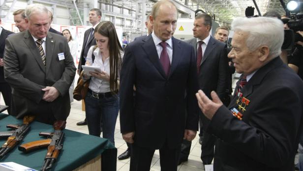 Wladimir Putin bei einem Treffen mit einem älteren Mann, der Orden trägt, während im Vordergrund Gewehre liegen.