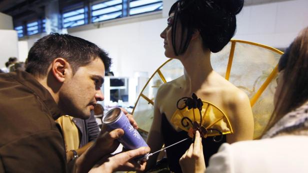 Ein Mann sprüht Haarspray auf das Kostüm einer Frau mit einem gelben Fächer und dunklen Verzierungen.