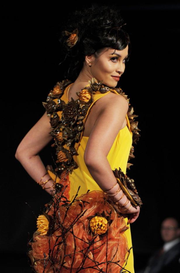 Eine Frau in einem gelben Kleid mit floralen Applikationen posiert auf einer dunklen Bühne.