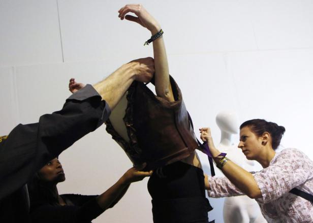 Mehrere Personen helfen, einer Schaufensterpuppe eine braune Lederweste anzuziehen.