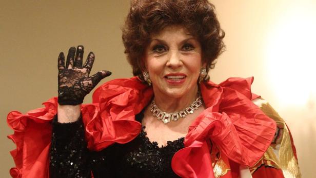 Eine ältere Frau mit rotem Rüschenkragen und Spitzenhandschuh winkt.