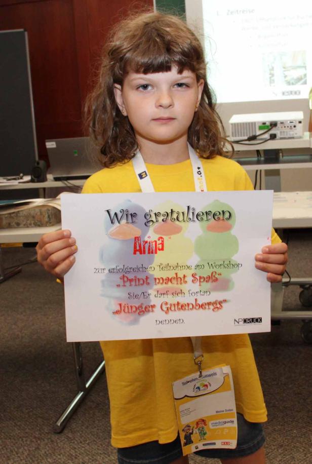 Ein junges Mädchen hält eine Urkunde mit der Aufschrift „Wir gratulieren Arina zur erfolgreichen Teilnahme am Workshop ‚Print macht Spaß‘. Sie/Er darf sich fortan ‚Jünger Gutenbergs‘ nennen.“.