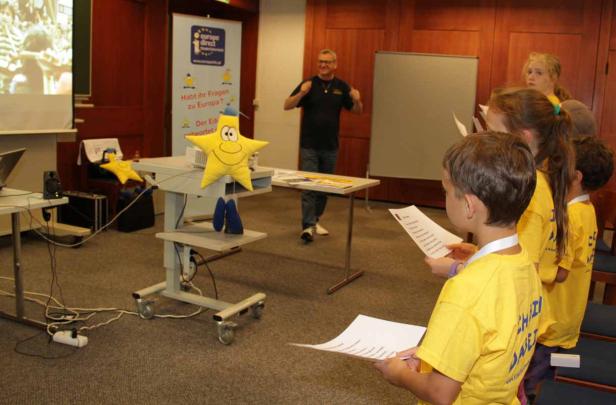 Kinder in gelben T-Shirts lesen vor einem Publikum in einem Raum mit „Europe Direct“-Werbung.