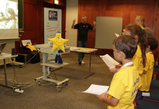 Kinder lesen in gelben T-Shirts vor einem Mann und einem Sternmaskottchen.
