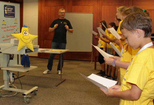 Eine Gruppe Kinder in gelben T-Shirts singt vor einem Mann und einem Europa-direct-Stand.