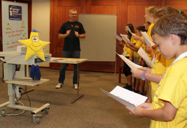 Eine Gruppe Kinder in gelben T-Shirts liest vor einem Mann und einem Europe Direct-Banner.