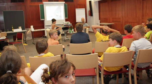 Kinder sitzen in einem Raum und schauen einer Präsentation zu.