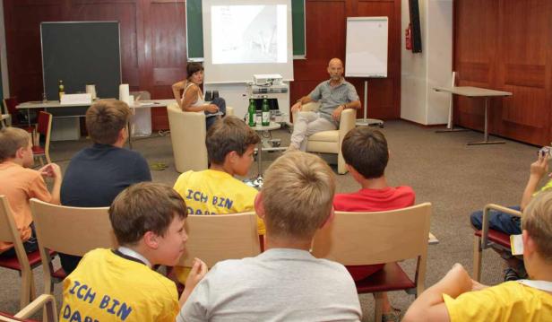 Eine Gruppe Kinder sitzt vor einem Mann und einer Frau, die eine Präsentation halten.