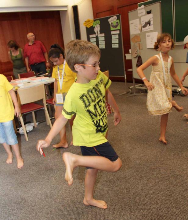 Barfüßige Kinder tanzen in einem Raum mit Tischen und Stühlen.