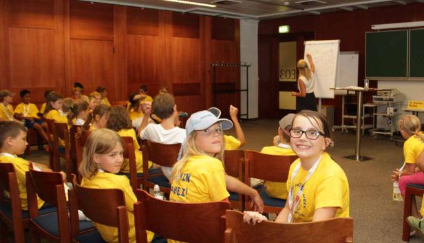 Eine Gruppe Kinder sitzt in einem Raum und hört einer Frau zu, die an einer Tafel schreibt.