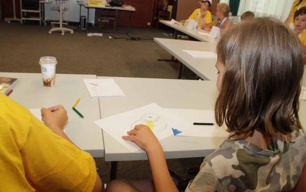Kinder sitzen an Tischen und malen Bilder mit Buntstiften.