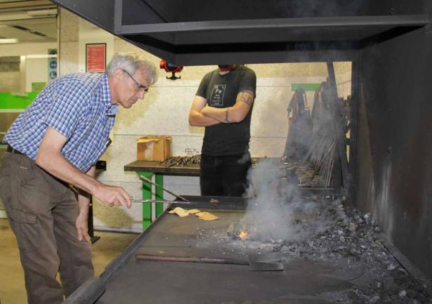 Ein Mann arbeitet an einer Schmiede, während ein anderer Mann zusieht.