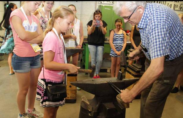Ein Mädchen schmiedet unter Anleitung eines Schmieds ein Stück Metall auf einem Amboss.