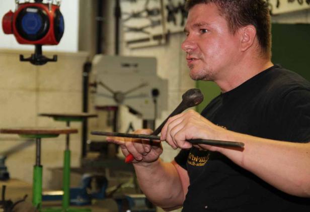 Ein Mann hält eine Metallstange und einen Hammer in einer Werkstatt.