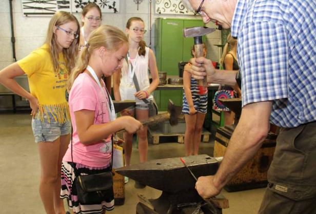 Ein Mädchen schmiedet unter Anleitung eines Schmieds ein Stück Metall auf einem Amboss.