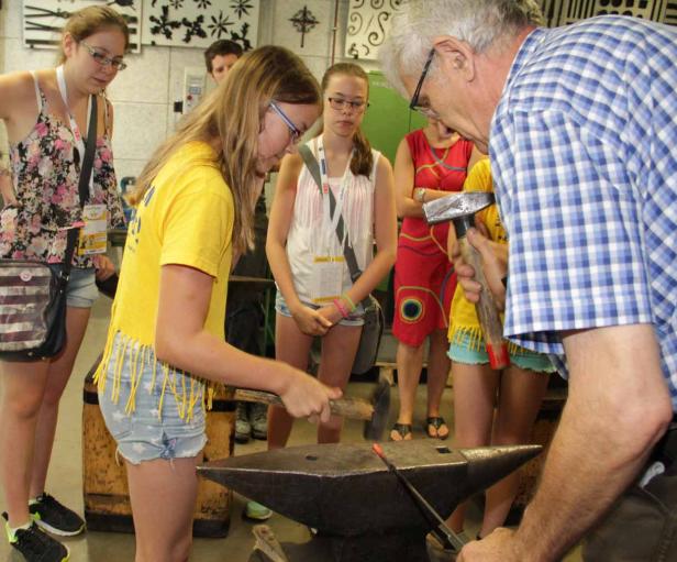 Ein Mädchen schmiedet unter Anleitung eines Mannes ein Stück Metall auf einem Amboss.