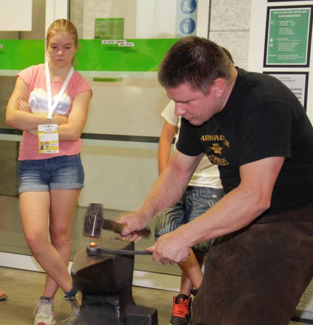 Ein Schmied bearbeitet glühendes Metall auf einem Amboss, während eine junge Frau zusieht.